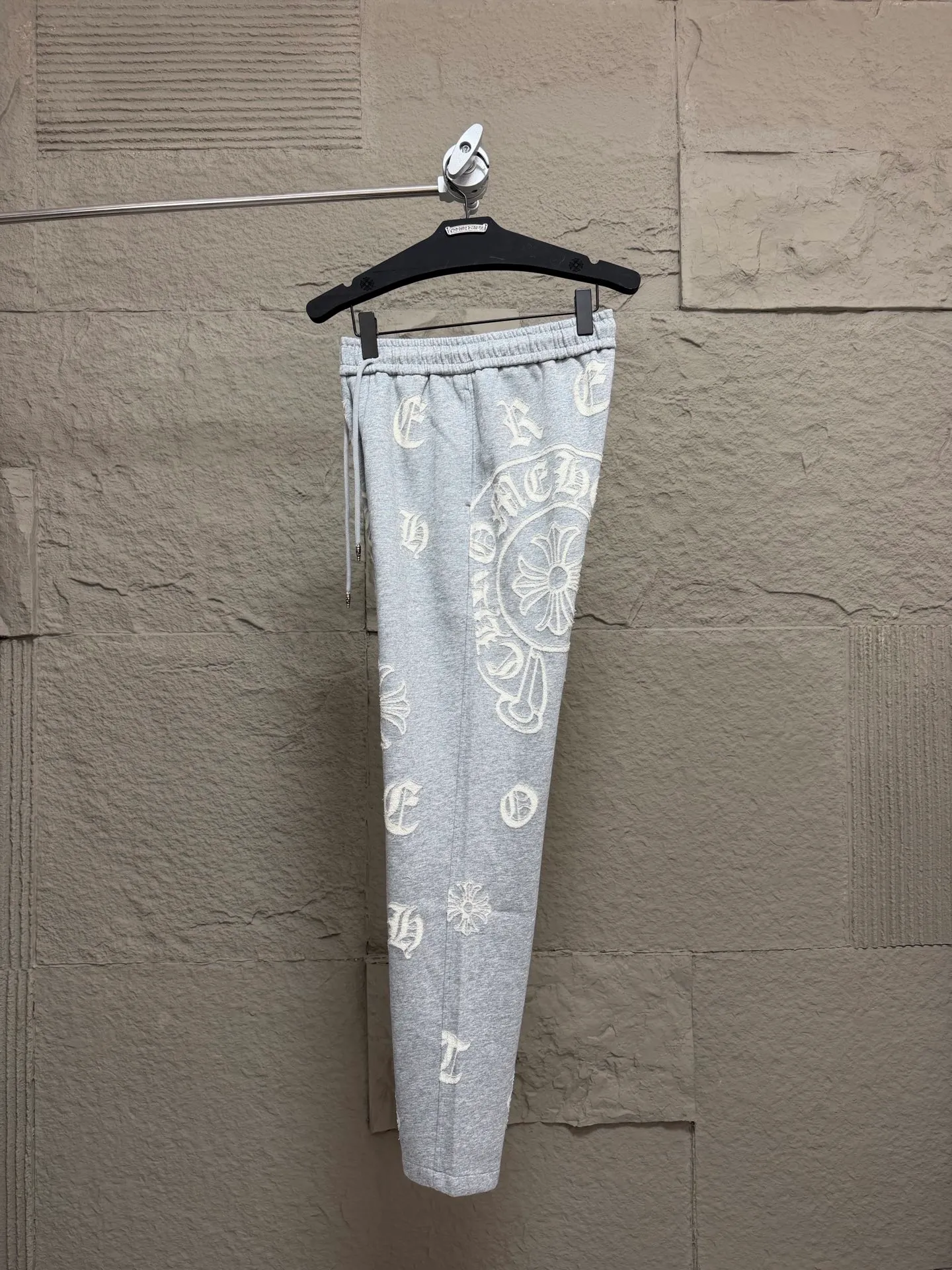 CH/Cro Heart 2026ss Burnt Flower Straight-leg Sweatpants Classic heavy-weight sweatpants with gray tones, Retro street style full-fitting, direct recognition, same style for men and women. ●Three-dimensional patchwork technology: The whole body is covered with Gothic letters + cross flower totems, using raw edge patchwork embroidery, with a mustache-like old texture, and uneven and layered patterns. It is not an ordinary print, and it is durable and not easy to fall off. ●Style tailoring: Loose, straight and wide-legged version, modifying the shape of the legs, hiding the flesh neatly, not flattering the figure, with an online drape, and the upper body has a relaxed street tone. ●Fabric and waist: Heavy knitted terry fabric, thick and soft, with good drape; elastic drawstring waist, with branded metal engraved rope ends, easy to put on and take off, and strong waist adaptability. ●Detailed design: The side pockets are convenient for carrying items, and the back pocket is decorated with cross-pattern patches. The front and rear patterns echo each other. Every detail is highly recognizable to the brand. ●Wearing scenarios: Suitable for street wear, daily outings, and trendy wear. Paired with sweaters, denim jackets, and leather jackets, you can easily create an American retro cool style. ●The overall gray base is paired with Cream White patches, which are low-key yet impactful. They are both comfortable and trendy. Versatile sweatpants, a must-have for fashionistas, are also available at the counter. SIZE：M L XL XXL XXXL